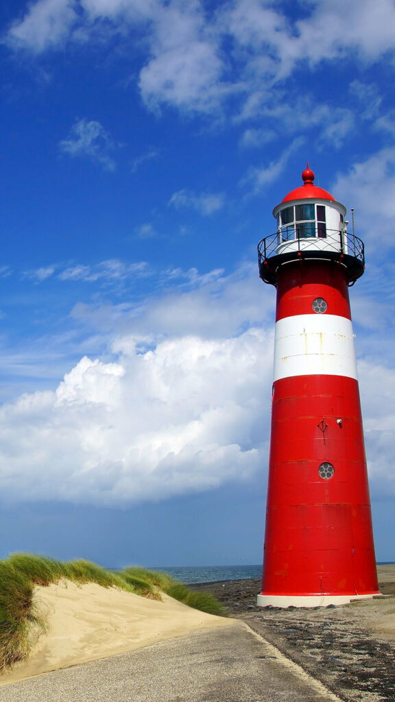 The image shows a red and white lighthouse standing tall near the shore. The lighthouse has a red top and two white bands around its middle. It is situated beside a sandy path with grassy dunes, leading towards a vast expanse of water. The sky is mostly clear with some scattered clouds, creating a bright and picturesque scene. The overall atmosphere is calm and serene, evoking a sense of coastal tranquility.